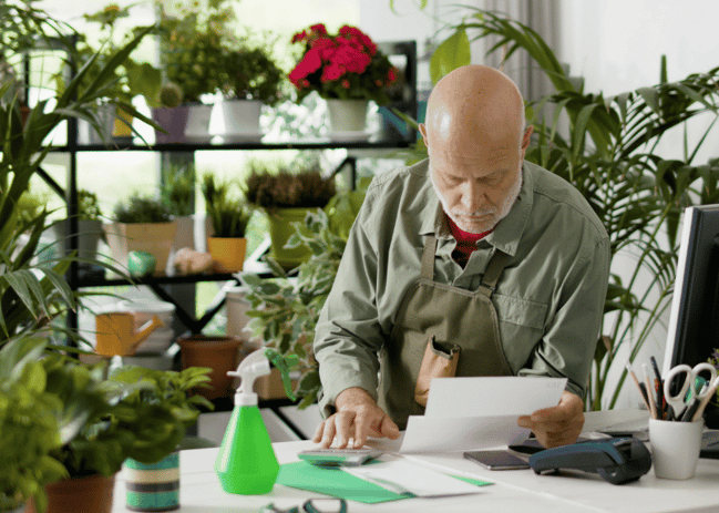 Older man looking at a paper while sitting inside his greenhouse. 