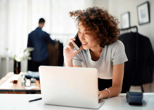 Woman leaning over desk with computer open and talking on her cellphone. 