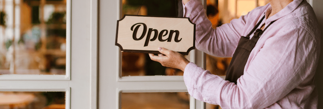 Older woman holding an open sign outside of her shop