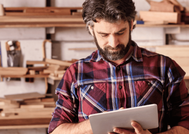 Middle aged man standing in his workshop looking at a tablet. 