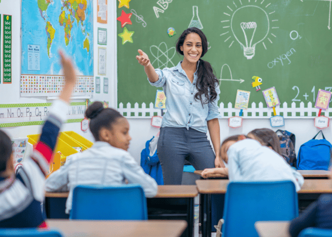 A female Hispanic teacher calling on a young student who is raising their hand. 