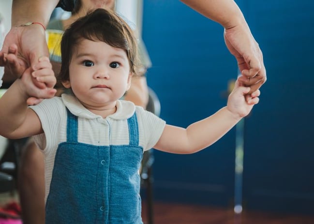 toddler girl taking first step while holding hands of parent (1)