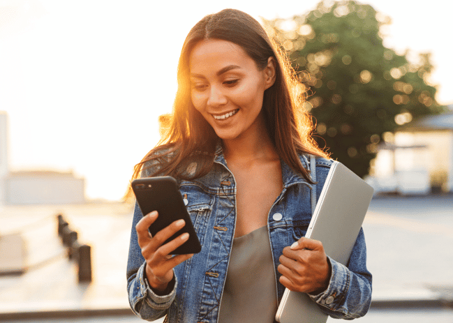 Woman looking down at her smartphone while holding a laptop in the other hand. 