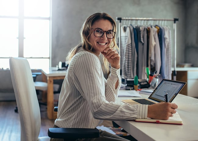 Business woman smiling at the camera while she has a computer in front of her and holds a pen