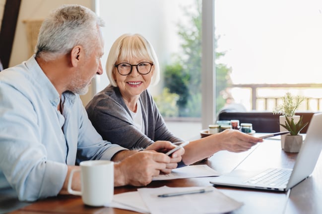 elderly couple at a desk with papers and pointing at their computer