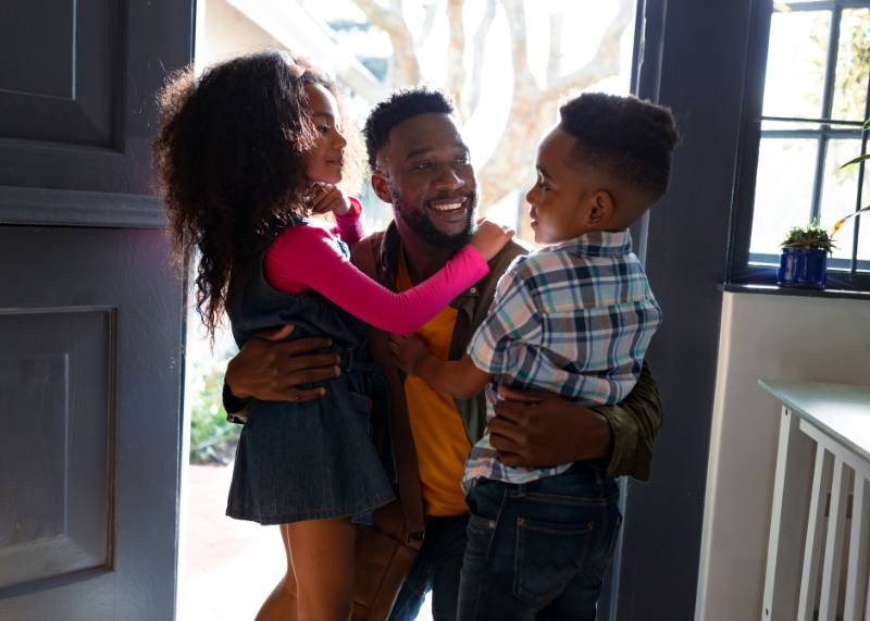 Man embracing his two small kids as he walks in the door.