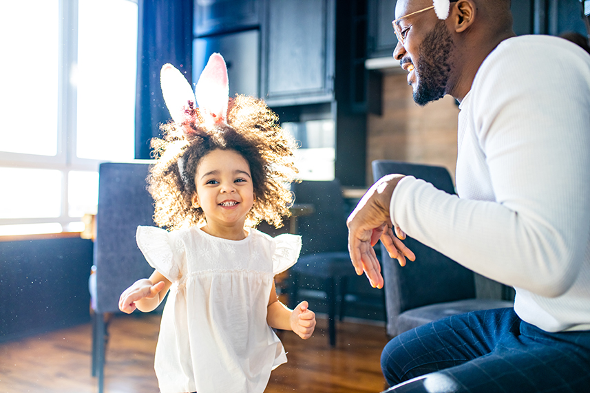 African American dad watching his little girl running across the room wearing bunny ears